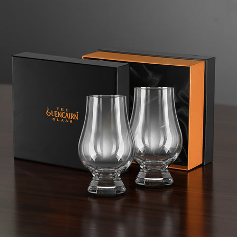 Two Glencairn whisky glasses in front of a branded box on a dark surface.