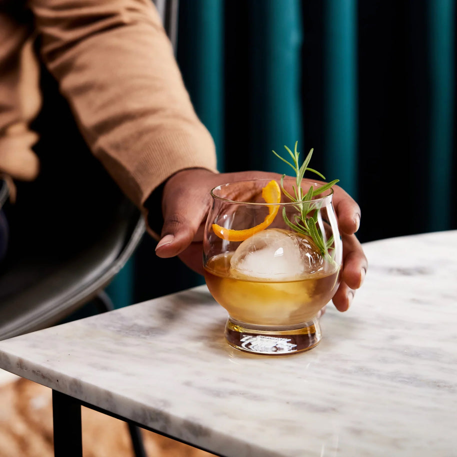 Whiskey glass with a round ice sphere, citrus twist, and herb garnish, creating a refined handcrafted cocktail moment