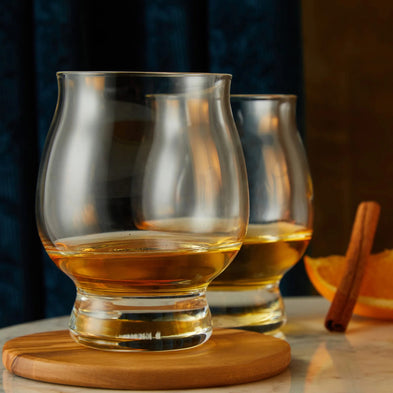 Two whiskey glasses with golden pours rest on wooden coasters, styled with orange and cinnamon for a warm, refined touch