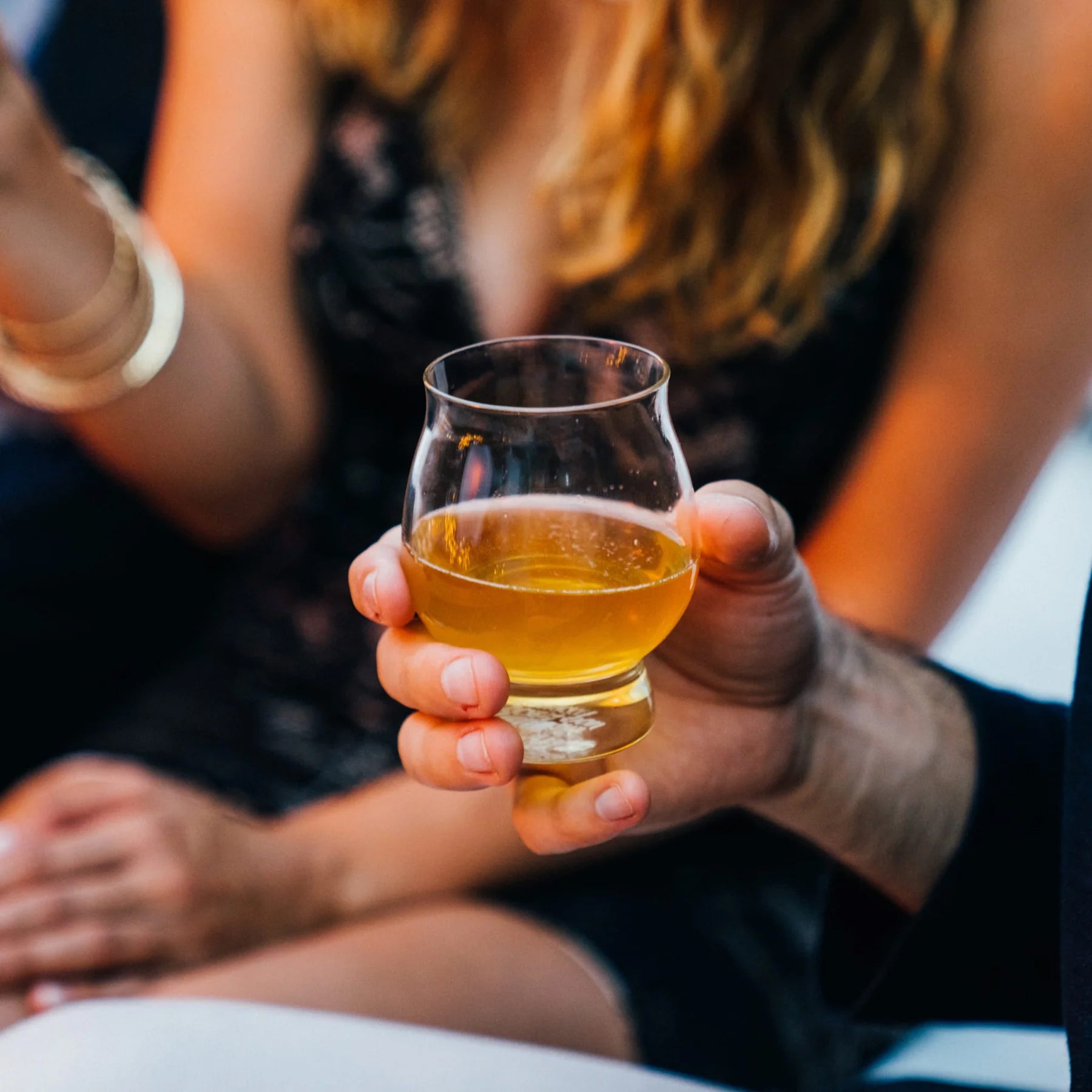 Hand holding a whiskey glass with golden liquid, capturing a warm, social moment in an intimate setting
