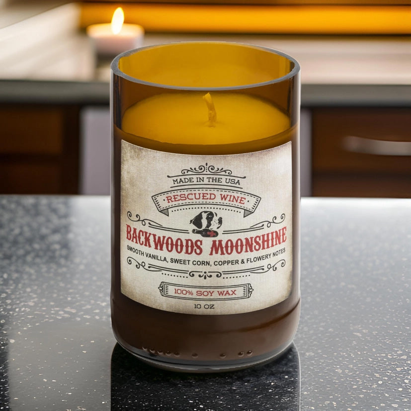 Brown glass candle labeled “Backwoods Moonshine.” The label reads: “Made in the USA. Rescued Wine. Backwoods Moonshine. Smooth vanilla, sweet corn, copper & flowery notes. 100% soy wax. 10 oz.” The candle wax inside is golden yellow, and it sits on a kitchen countertop
