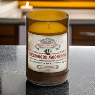 Brown glass candle labeled “Backwoods Moonshine.” The label reads: “Made in the USA. Rescued Wine. Backwoods Moonshine. Smooth vanilla, sweet corn, copper & flowery notes. 100% soy wax. 10 oz.” The candle wax inside is golden yellow, and it sits on a kitchen countertop