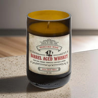 Brown glass candle labeled “Barrel Aged Whiskey.” The label reads: “Made in the USA. Rescued Wine. Barrel Aged Whiskey. Aged oak, cedar, tobacco, leather & herbs. 100% soy wax. 10 oz.” The candle wax inside is golden yellow, and it rests on a speckled countertop 
