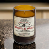 Brown glass candle labeled “Bourbon and Tobacco.” The label reads: “Made in the USA. Rescued Wine. Bourbon and Tobacco. Caramel, oak, herbs & tobacco. 100% soy wax. 10 oz.” The candle wax inside is golden yellow, and it sits on a polished countertop
