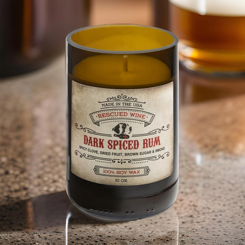 Brown glass candle labeled “Dark Spiced Rum.” The label reads: “Made in the USA. Rescued Wine. Dark Spiced Rum. Spicy clove, dried fruit, brown sugar & smoke. 100% soy wax. 10 oz.” The candle wax is golden yellow, and the candle sits on a polished countertop 