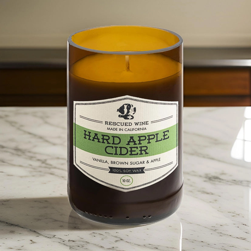 Brown glass candle with a cream and green label reading “Rescued Wine – Hard Apple Cider.” The scent notes listed are vanilla, brown sugar, and apple. The candle sits on a white marble countertop