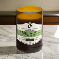 Brown glass candle with a cream and green label reading “Rescued Wine – Hard Apple Cider.” The scent notes listed are vanilla, brown sugar, and apple. The candle sits on a white marble countertop