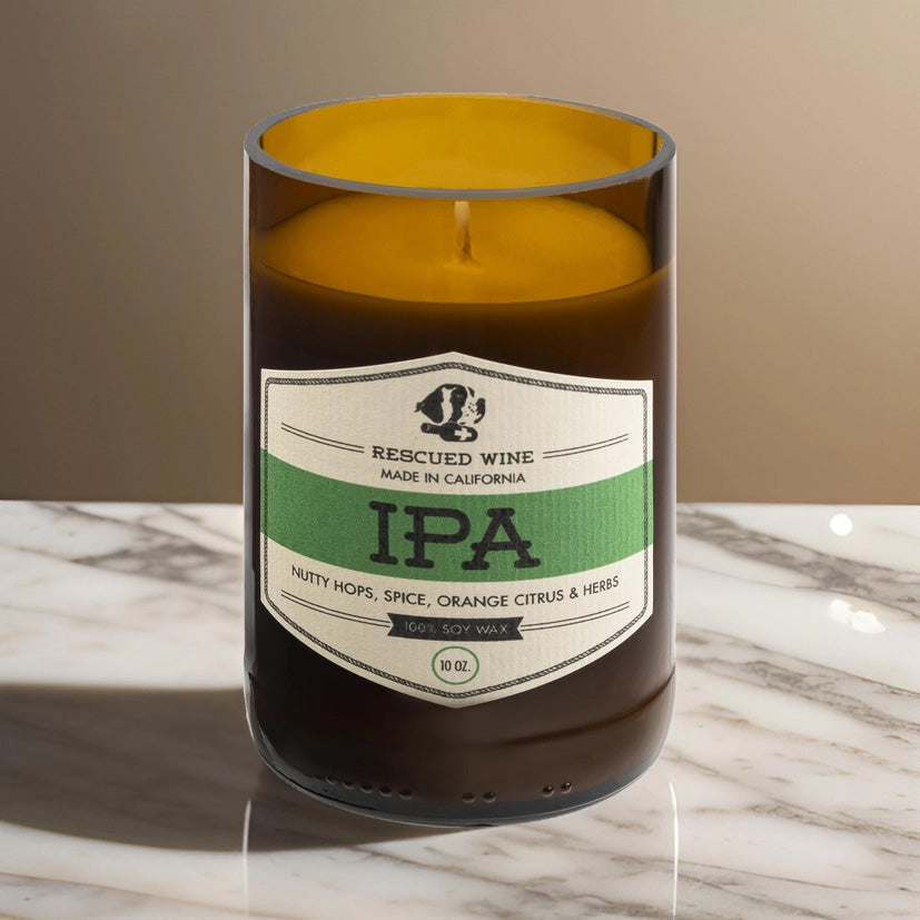 Brown glass candle with a cream and green label reading “Rescued Wine – IPA.” The scent notes listed are nutty hops, spice, orange citrus, and herbs. The candle sits on a marble surface