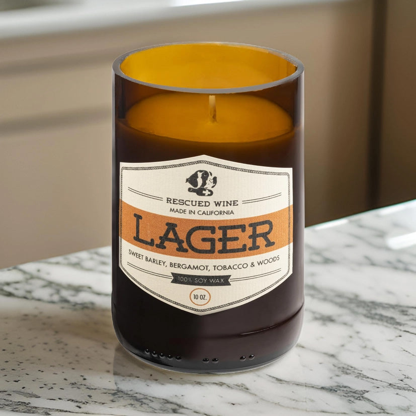 Brown glass candle with a cream and orange label reading “Rescued Wine – Lager.” The scent notes listed are sweet barley, bergamot, tobacco, and woods. The candle is placed on a white marble countertop