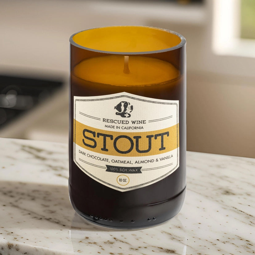 Brown glass candle with a white and gold label reading “Rescued Wine – Stout.” The scent notes listed are dark chocolate, oatmeal, almond, and vanilla. The candle sits on a marble countertop