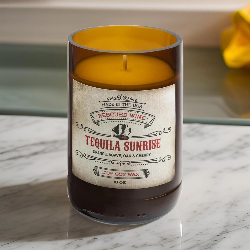 Brown glass candle labeled “Tequila Sunrise.” The label reads: “Made in the USA. Rescued Wine. Tequila Sunrise. Orange, agave, oak & cherry. 100% soy wax. 10 oz.” The candle wax inside is golden yellow, and it sits on a white marble surface with a blurred yellow cloth in the background