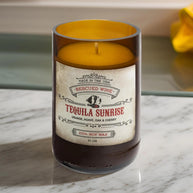 Brown glass candle labeled “Tequila Sunrise.” The label reads: “Made in the USA. Rescued Wine. Tequila Sunrise. Orange, agave, oak & cherry. 100% soy wax. 10 oz.” The candle wax inside is golden yellow, and it sits on a white marble surface with a blurred yellow cloth in the background