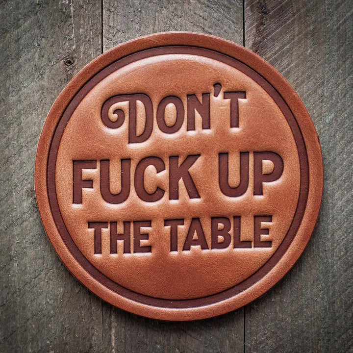 Round leather coaster embossed with the phrase ‘Don’t Fuck Up The Table’ in bold block lettering, rustic novelty barware