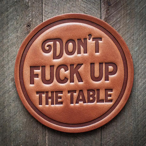 Round leather coaster embossed with the phrase ‘Don’t Fuck Up The Table’ in bold block lettering, rustic novelty barware