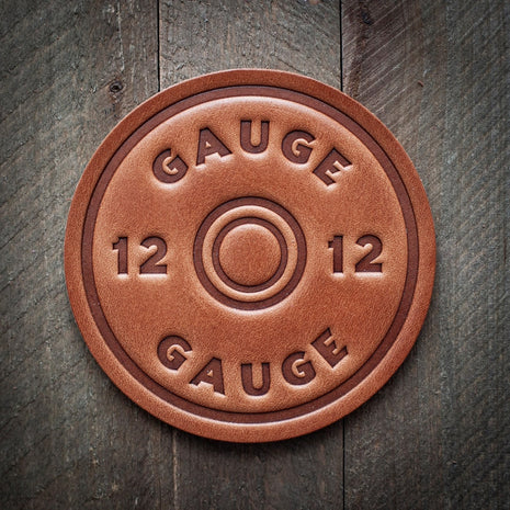 Round leather coaster designed to resemble a 12-gauge shotgun shell headstamp, embossed with the text ‘12 Gauge’ around a center ring