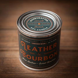 Sugarhouse Leather & Bourbon soy candle in 8oz metal tin. Dark green and orange vintage-style label with text: Handcrafted small batch, hand poured, cotton wick, 40 hour burn. Rustic wood table background