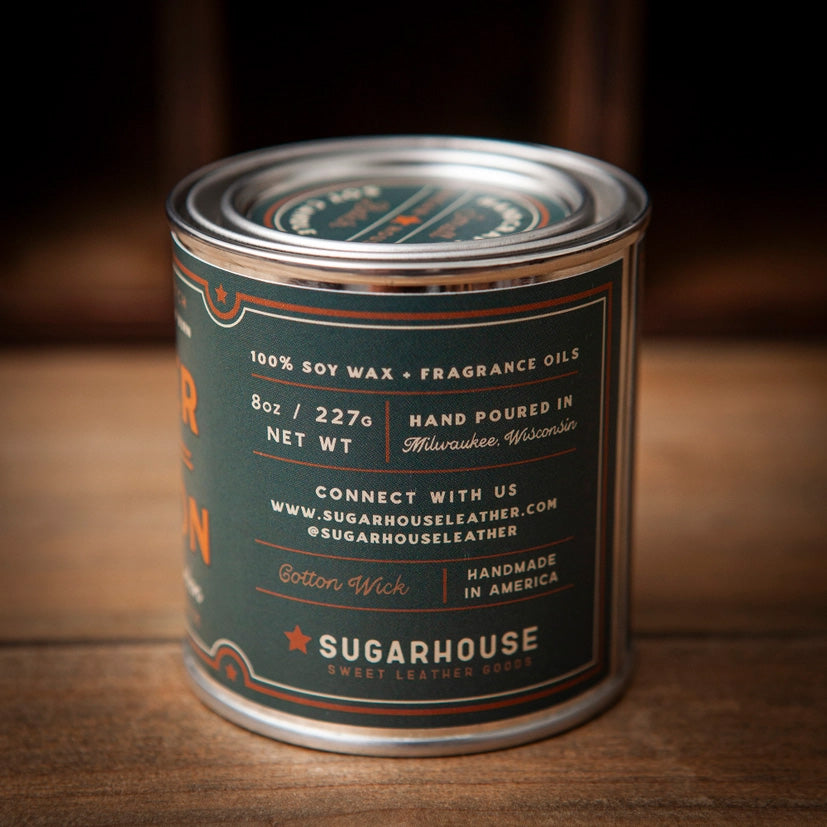 Sugarhouse Sweet Leather Goods candle in an 8oz metal tin with dark green and orange label. Text highlights: 100% soy wax, fragrance oils, hand-poured in Milwaukee, Wisconsin, cotton wick, handmade in America. Rustic wood background