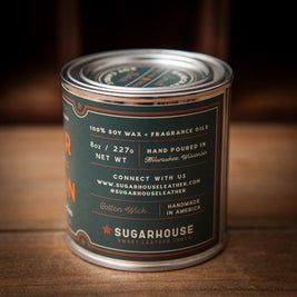 Sugarhouse Sweet Leather Goods candle in an 8oz metal tin with dark green and orange label. Text highlights: 100% soy wax, fragrance oils, hand-poured in Milwaukee, Wisconsin, cotton wick, handmade in America. Rustic wood background