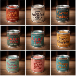 Collection of nine vintage-inspired soy candles in tin cans, each with bold, retro-style typography. Labels read: “Call Me Old Fashioned,” “Light a Fucking Candle,” “Leather & Bourbon,” “Cabin Fever,” “Lake Time,” “Original Cowboy,” “Unicorn Breath,” “Sexy Time,” and “Happy Place.” The candles feature colorful designs in shades of red, cream, teal, pink, and yellow, all hand-crafted by Sugarhouse.