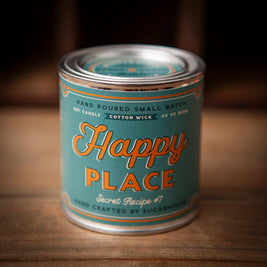 Sugarhouse candle tin with teal label and silver lid. The label features the text “Happy Place” in large orange script, with smaller text reading “Secret Recipe #7,” “Hand Poured Small Batch Soy Candle,” “Cotton Wick,” and “40 hr burn.” Decorative orange accents frame the design.
