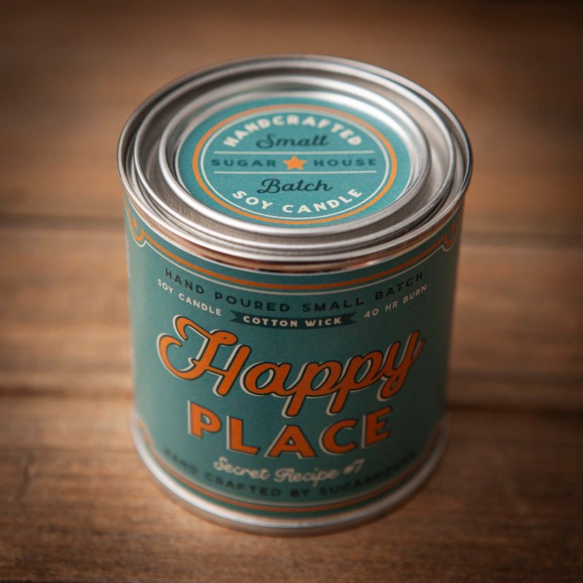 Sugarhouse candle tin with teal label and silver lid. The label reads “Happy Place” in large orange script, with “Secret Recipe #7,” “Hand Poured Small Batch Soy Candle,” “Cotton Wick,” and “40 hr burn.” The lid features the Sugarhouse logo with text “Handcrafted Small Batch Soy Candle.”