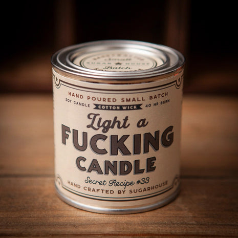 Sugarhouse candle tin with a beige label that reads “Light a Fucking Candle” in bold black lettering. Additional text includes “Hand Poured Small Batch,” “Soy Candle,” “Cotton Wick, 40-Hour Burn, and “Secret Recipe #33"