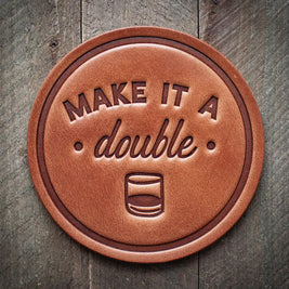 Round leather coaster embossed with the phrase ‘Make It a Double’ and a whiskey glass icon, rustic barware style.