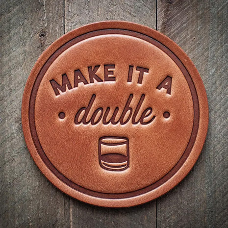 Round leather coaster embossed with the phrase ‘Make It a Double’ and a whiskey glass icon, rustic barware style.