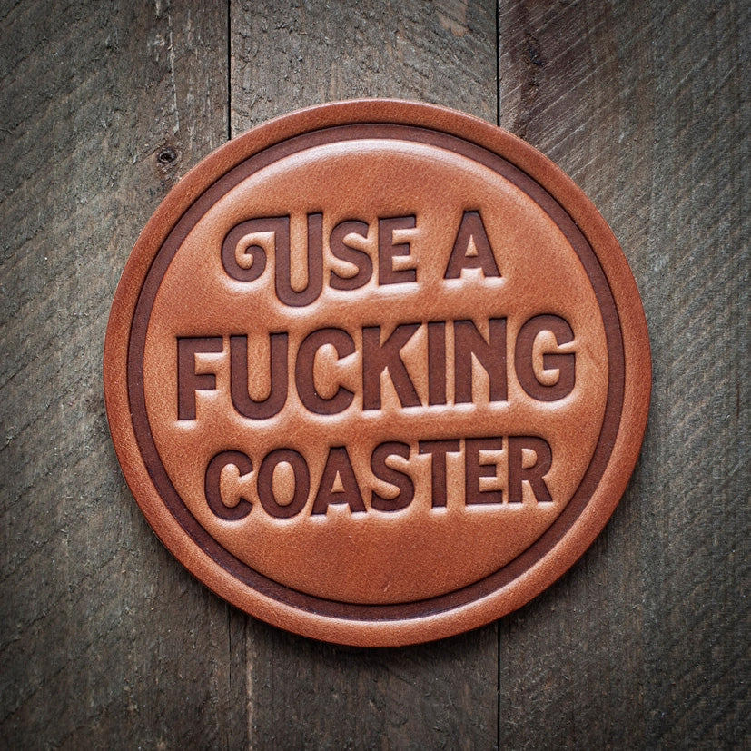 Round leather coaster embossed with the phrase ‘USE A FUCKING COASTER’ in bold capital letters, with a rustic background