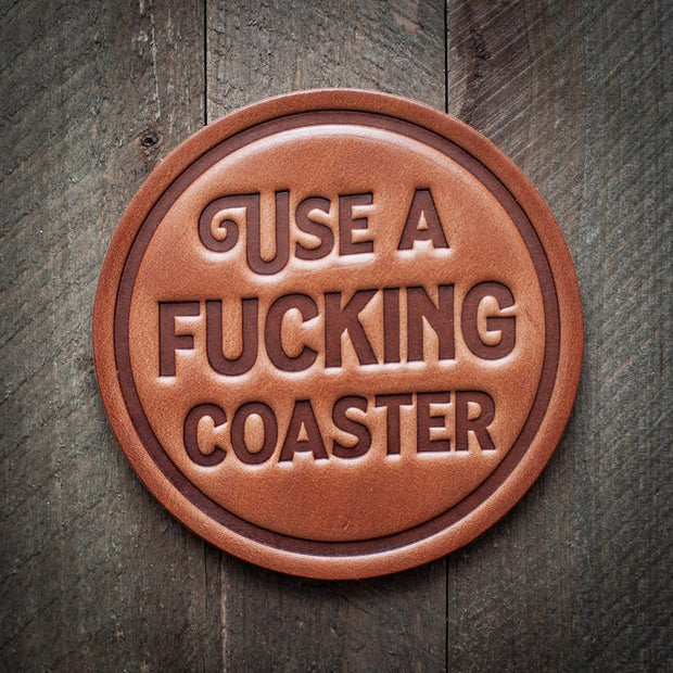 Round leather coaster embossed with the phrase ‘USE A FUCKING COASTER’ in bold capital letters, with a rustic background